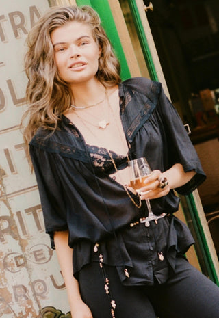 a-woman-wearing-a-black-sheer-lace-trim-blouse-and-black-pants-holding-a-wine-glass-in-front-of-a-rustic-doorway