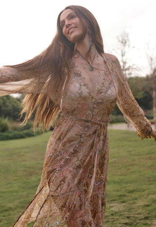 a-woman-outdoors-in-a-grassy-park-wearing-a-flowing-sheer-boho-style-maxi-dress-with-an-intricate-paisley-and-floral-pattern-in-warm-beige-brown-and-muted-pink-tones-she-is-captured-mid-spin-with-her-hair-blowing-wearing-layered-necklaces