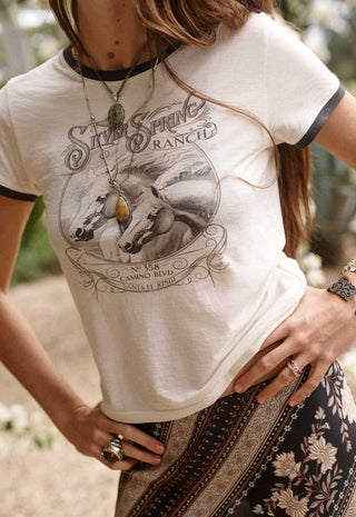 A woman wearing a cream-colored ringer t-shirt with black trim and a vintage 'Silk Spring Ranch' graphic print on the front